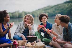 A group of young friends having fun on picnic near a lake, sitting on blanket and eating watermelon.