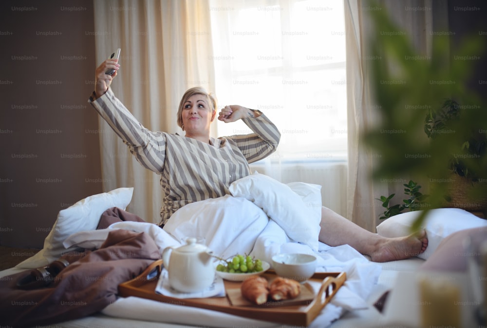 Eine glückliche übergewichtige Frau mit Selfie beim Frühstück im Bett zu Hause.