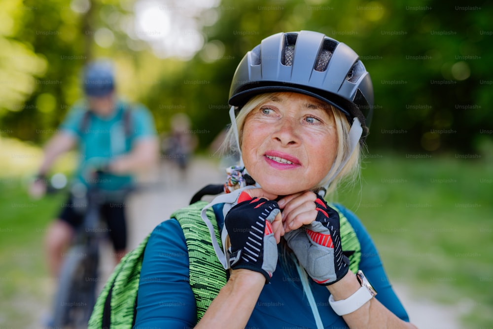 Um casal sênior ativo andando de bicicleta no parque de verão, a mulher está colocando capacete, conceito de estilo de vida saudável.