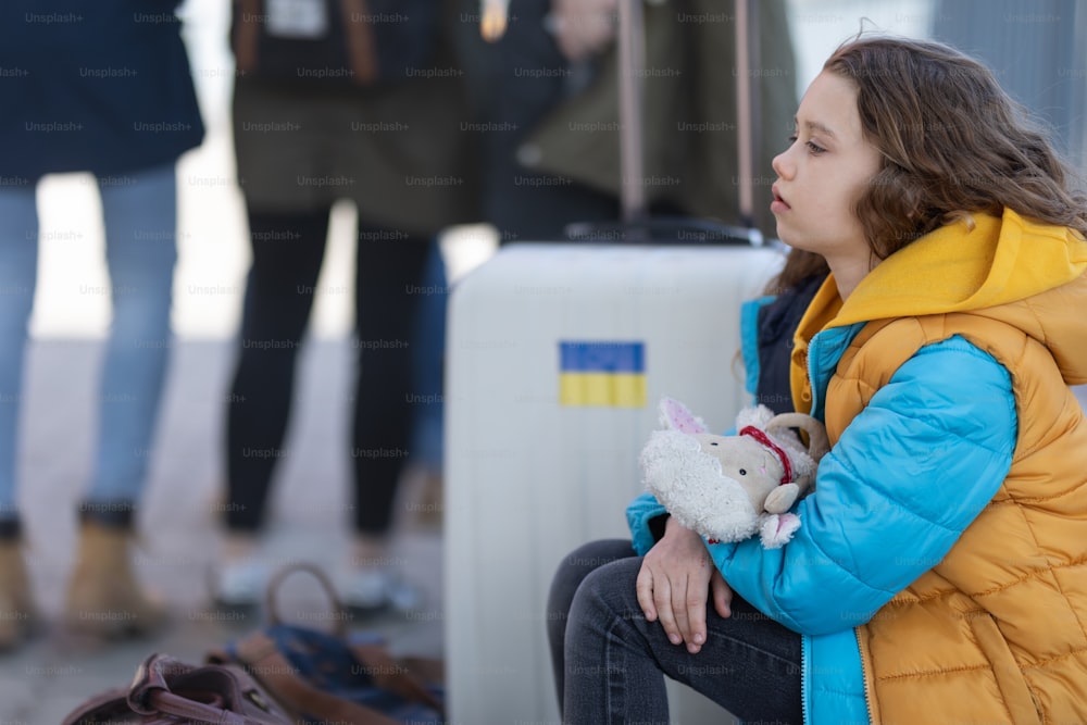 A sad Ukrainian immigrant child with luggage waiting at train station, Ukrainian war concept.