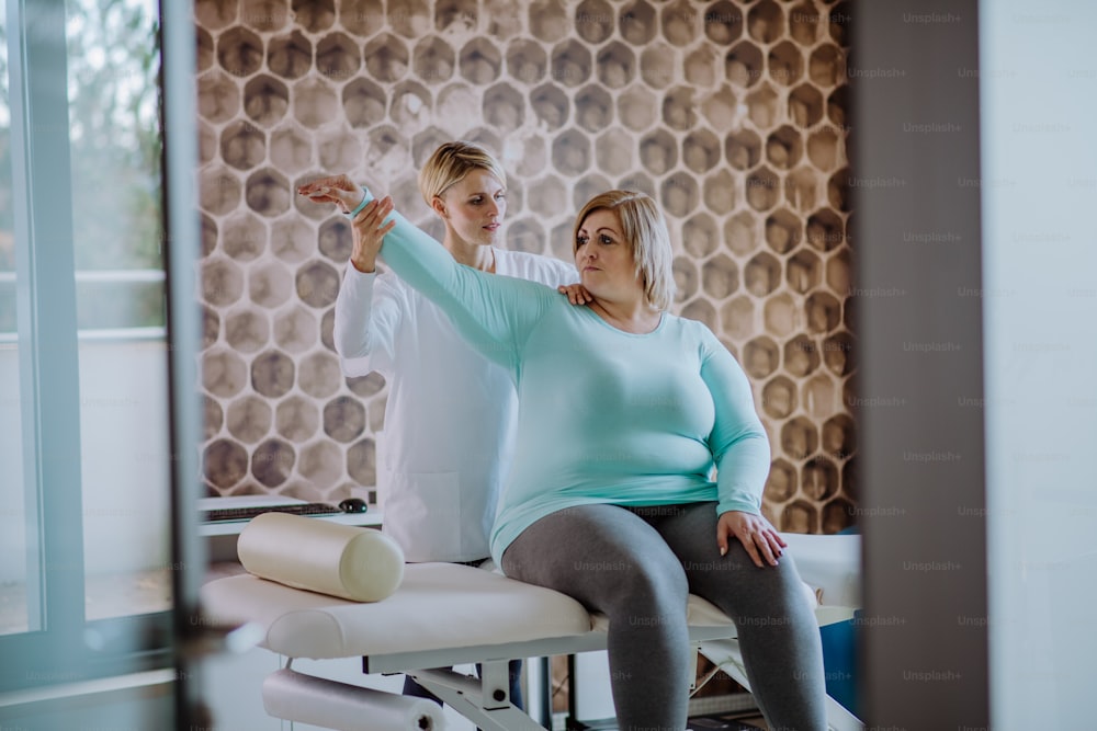 Una mujer fisioterapeuta adulta media que hace ejercicio con una mujer con sobrepeso en el interior de un centro de rehabilitación