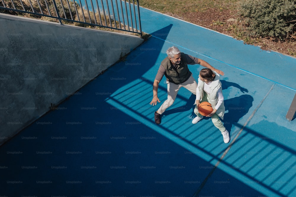 A father and teenage daughter playing basketball outside at court, high angle view.