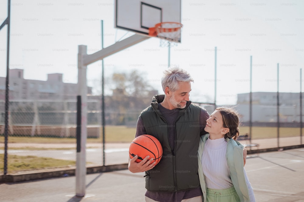 A happy father and teen daughter embracing and looking at camera outside at basketball court.