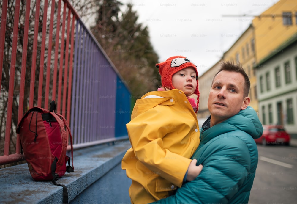 Ein Vater, der seine kleine Tochter mit Down-Syndrom zur Schule bringt, draußen auf der Straße.