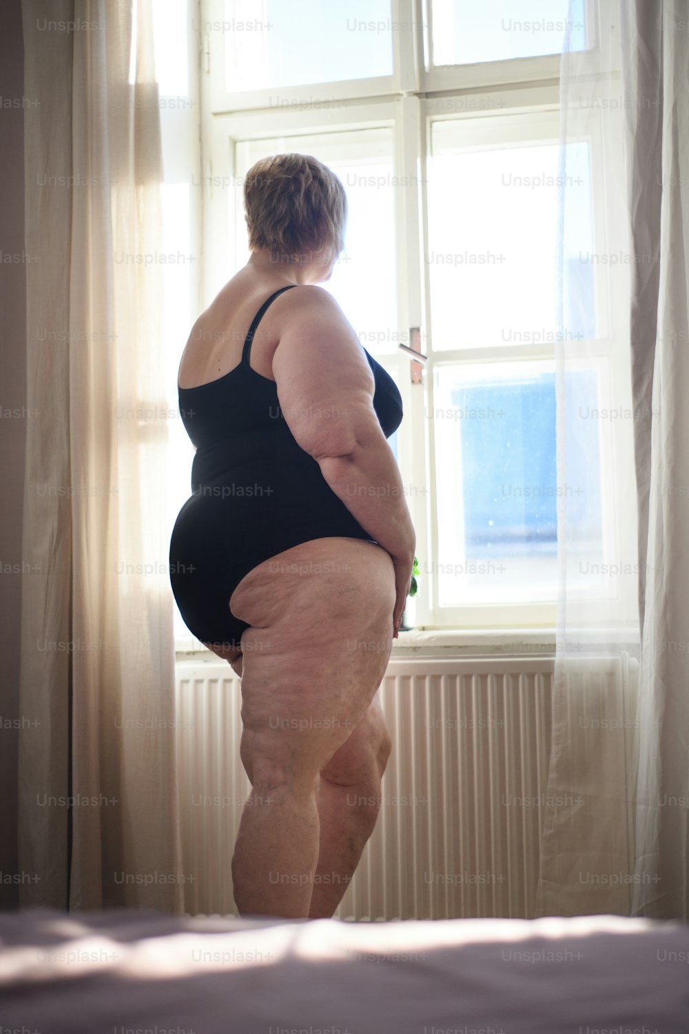 A rear view of lonely fat woman standing and looking through the window at home.