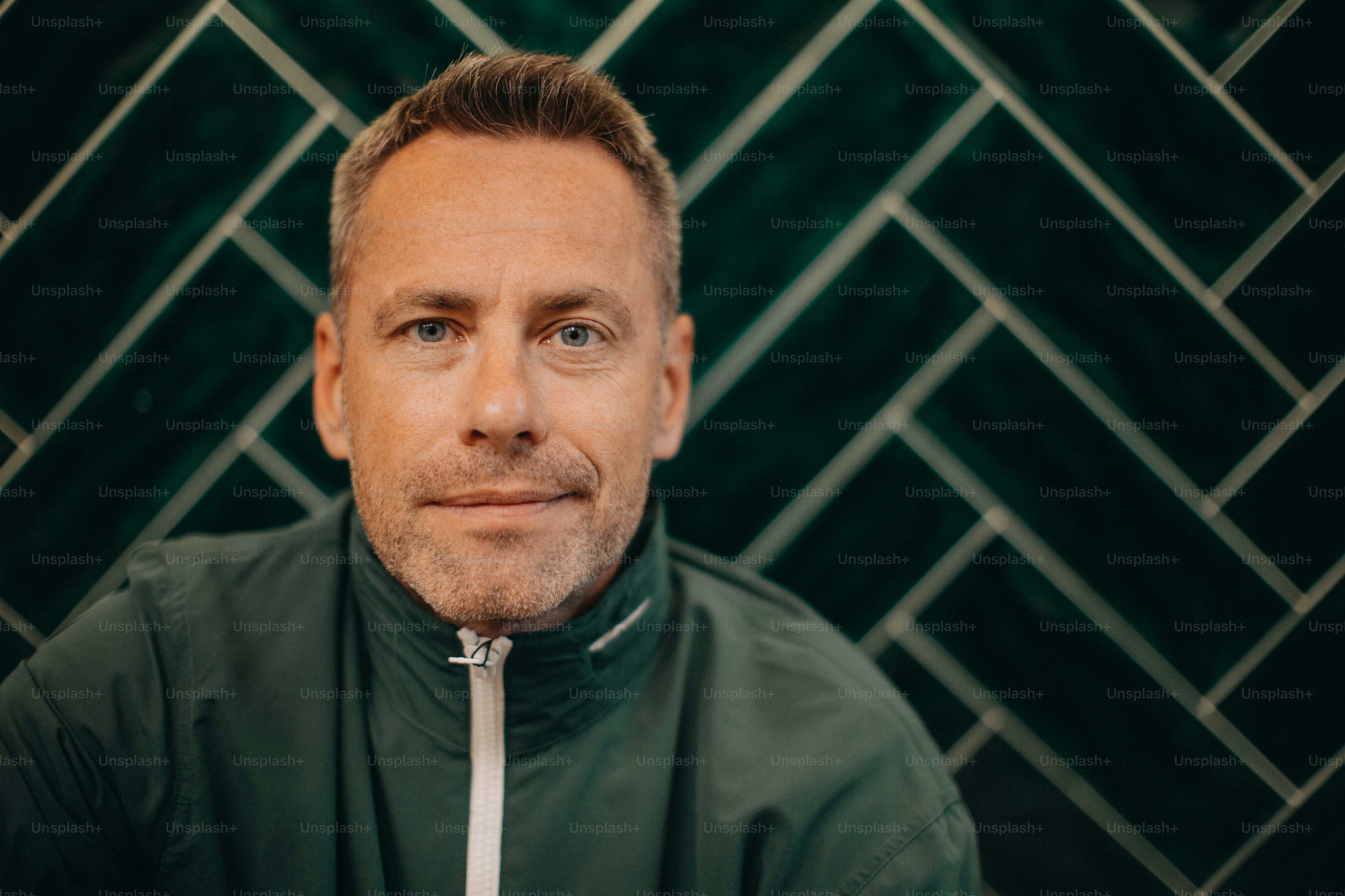 A portrait of middle-aged man in jacket sitting agiant dark tiled wall.