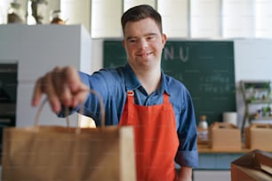 A cheerful young Down Syndrome waiter holding take away order in restaurant, social inclusion concept.