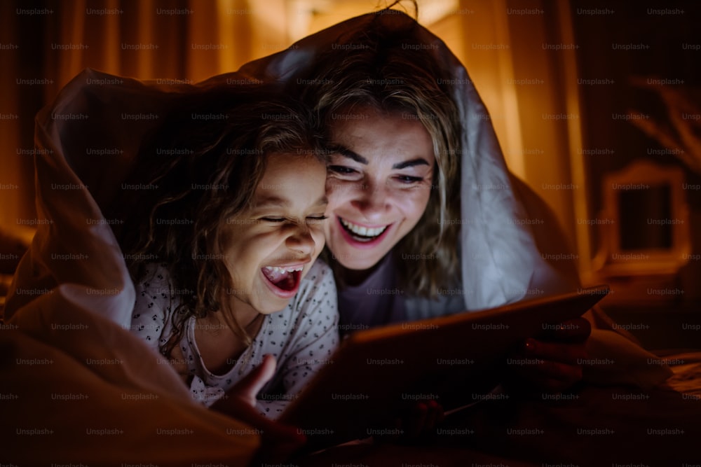 Eine glückliche Mutter mit ihrer kleinen Tochter, die unter der Decke liegt und zu Hause Filme auf dem Tablet schaut.