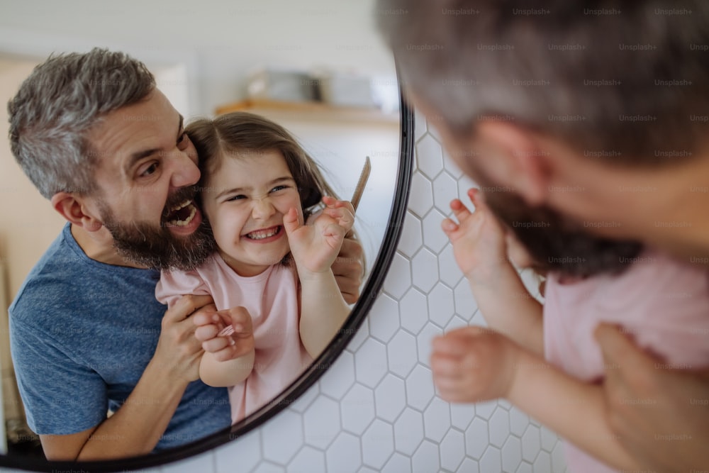 A father having fun with his little daughter, making grimaces in mirror in bathroom at home.
