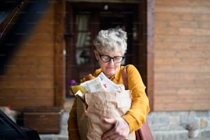 Senior woman with shopping in paper bag standing outdoors by car.