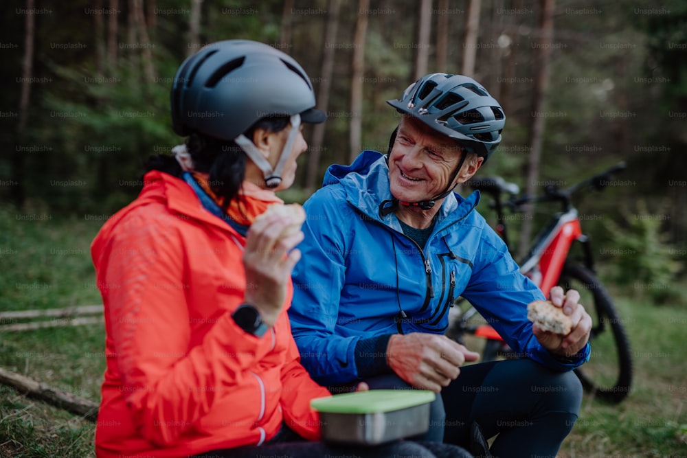 Una feliz pareja de ciclistas mayores con comer bocadillos al aire libre en el bosque en un día de otoño.