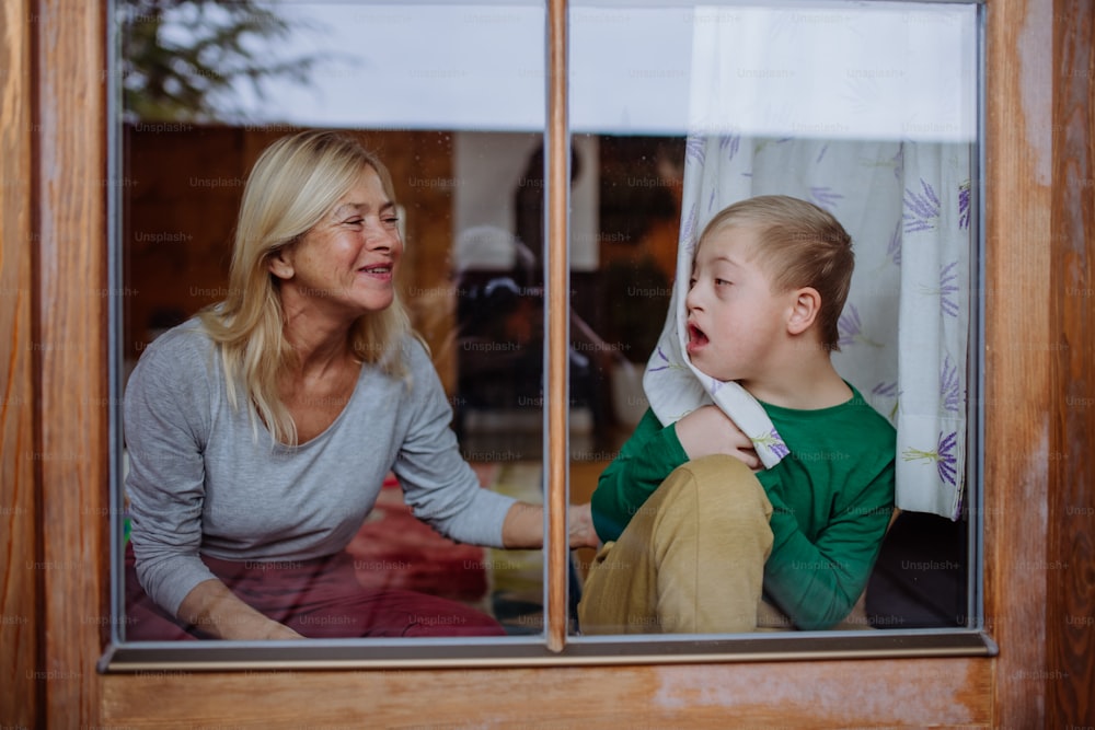 Um menino com síndrome de Down com sua avó olhando para a câmera através da janela em casa.