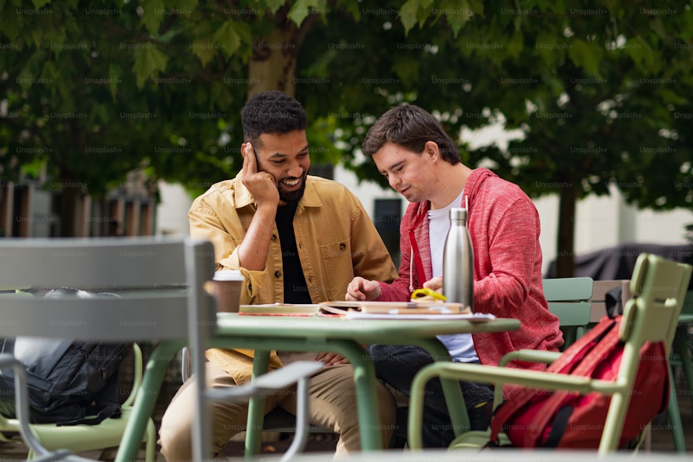 Un giovane con la sindrome di Down con il suo amico mentore seduto all'aperto in un caffè e studiando.