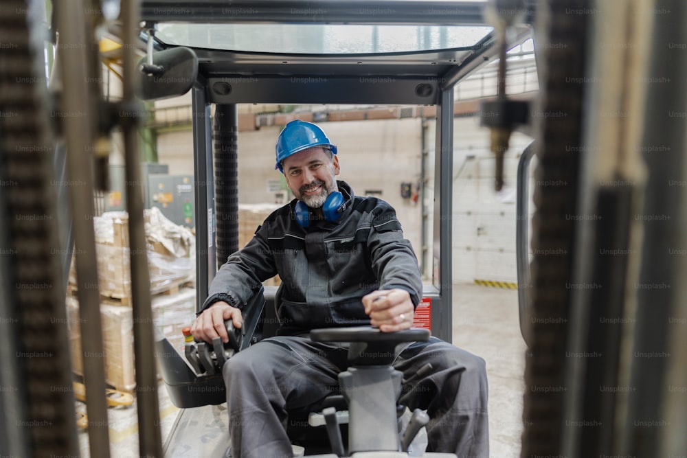 A happy mature man fork lift truck driver lifting pallet in storage warehouse and looking at camera.