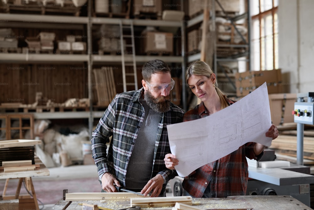 Two carpenters man and woman looking at blueprints indoors in a carpentery workshop.