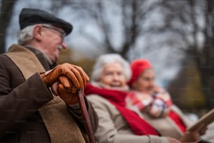 A group of happy senior friends sitting on bench in town park in autumn, focus on man's hands in gloves