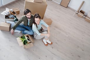 A high angle view of young couple with boxes sitting on floor when moving in new flat, new home and relocation concept.