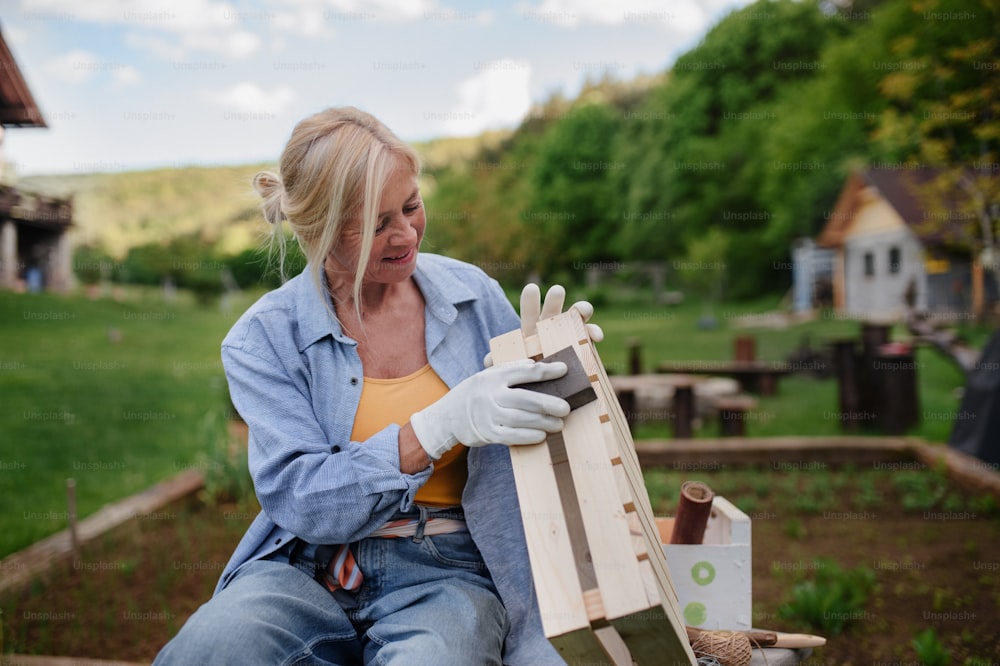 A happy senior woman renovating wooden crate outdoors in garden.