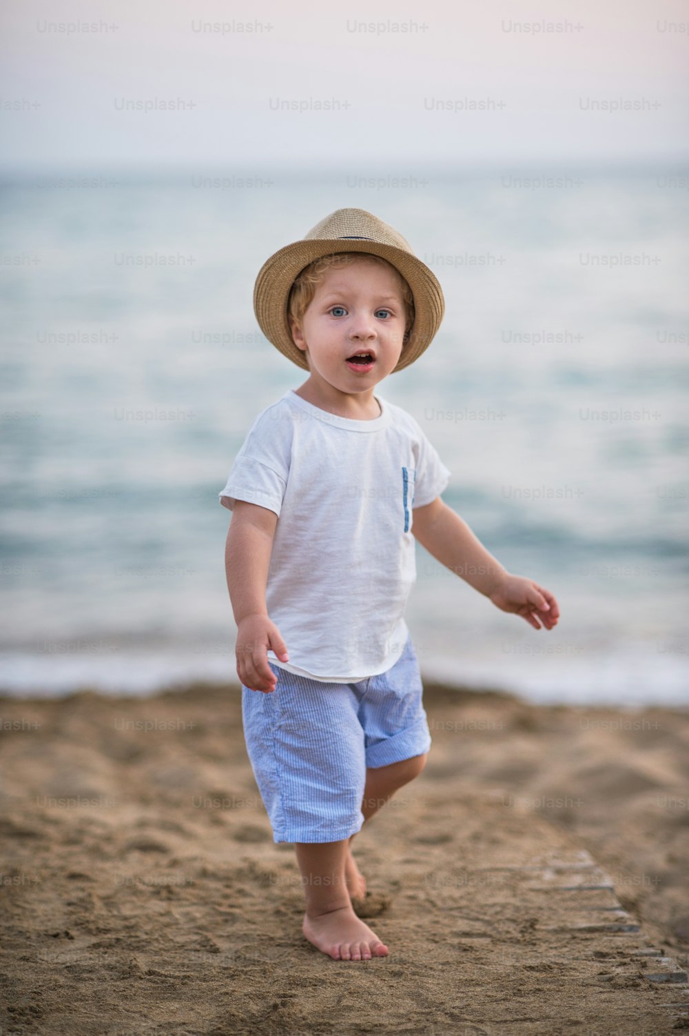A portrait of small blond toddler boy with hat standing on beach on summer holiday.