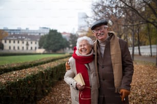 A happy senior couple on walk outdoors in park in autumn, embracing, laughing and looking at camera