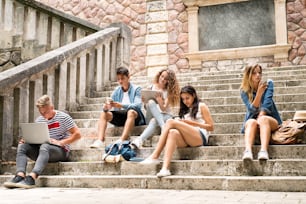 Group of attractive teenage students sitting on stone steps in front of university holding tablet, laptop and smart phones, reading or watching something.