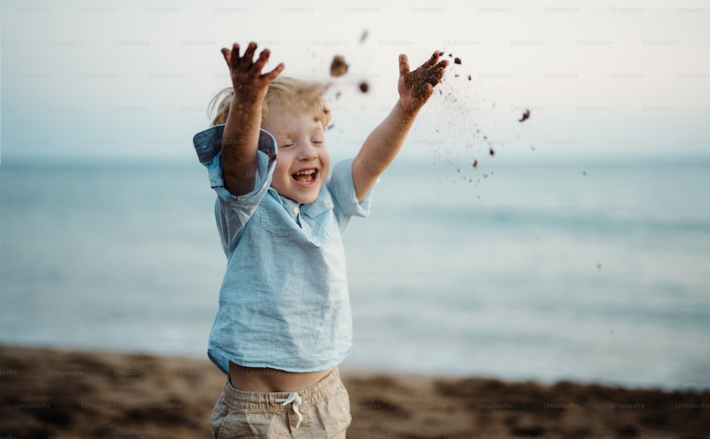 A cheerful small toddler boy standing on beach on summer holiday, throwing sand.