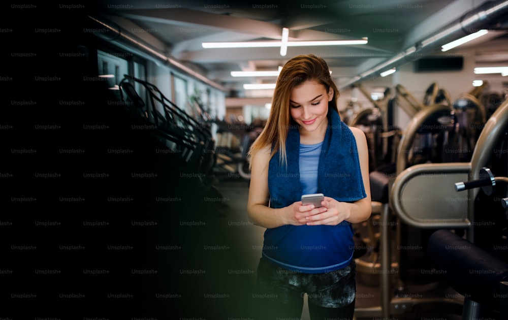 A portrait of happy young girl or woman in a gym, using smartphone.