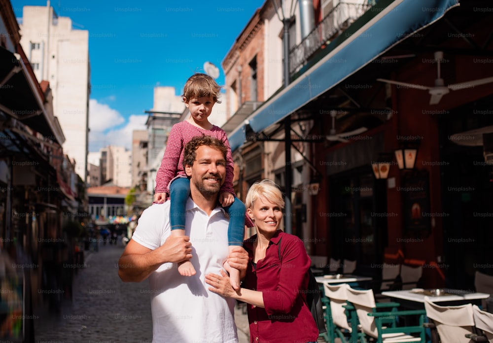 Young family with small daughter standing outdoors in town on holiday, giving piggyback ride.