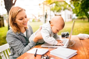 Beautiful young woman with a baby studying outdoors. A mother with laptop in the garden.
