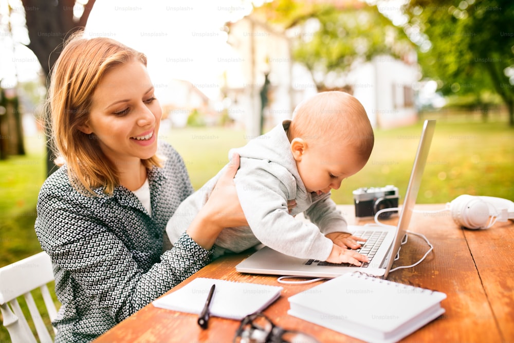 Beautiful young woman with a baby studying outdoors. A mother with laptop in the garden.