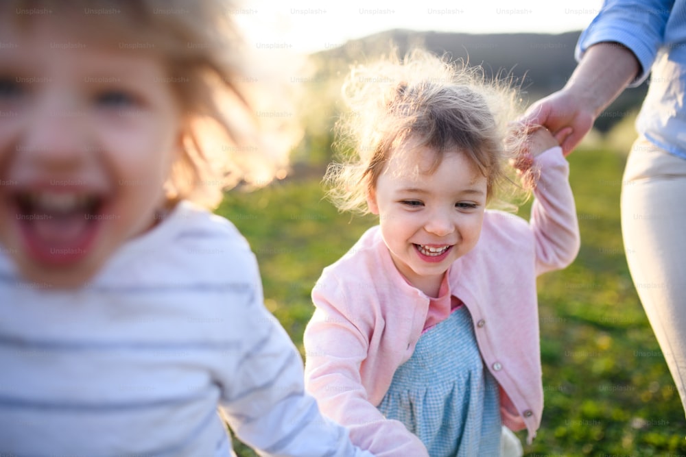 Duas crianças pequenas com mãe irreconhecível correndo ao ar livre em pomar na primavera.