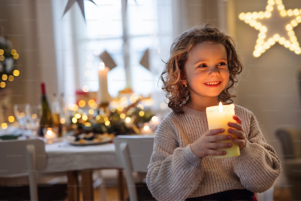 Vista frontal de una niña alegre de pie en el interior en Navidad, sosteniendo una vela.