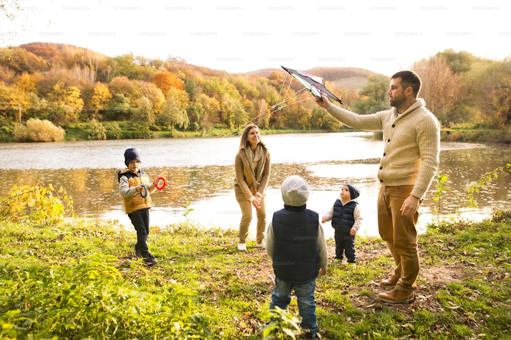 Beautiful young family on a walk at the lake in forest. Mother and father with their three sons with kite outside in colorful autumn nature.