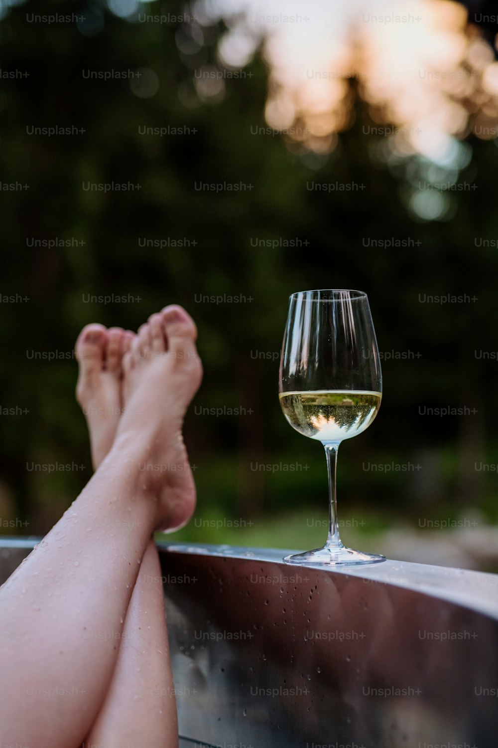 Una joven irreconocible con los pies en alto relajándose con una copa de vino en la bañera de hidromasaje al aire libre en la naturaleza.
