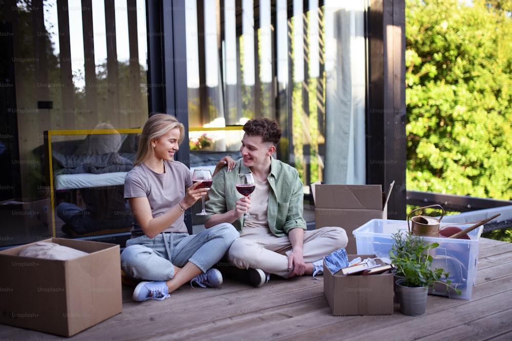 A cheerful young couple moving in their new tiny house in woods sitting on floor on terrace and drinking wine. Conception of moving and sustainable living.