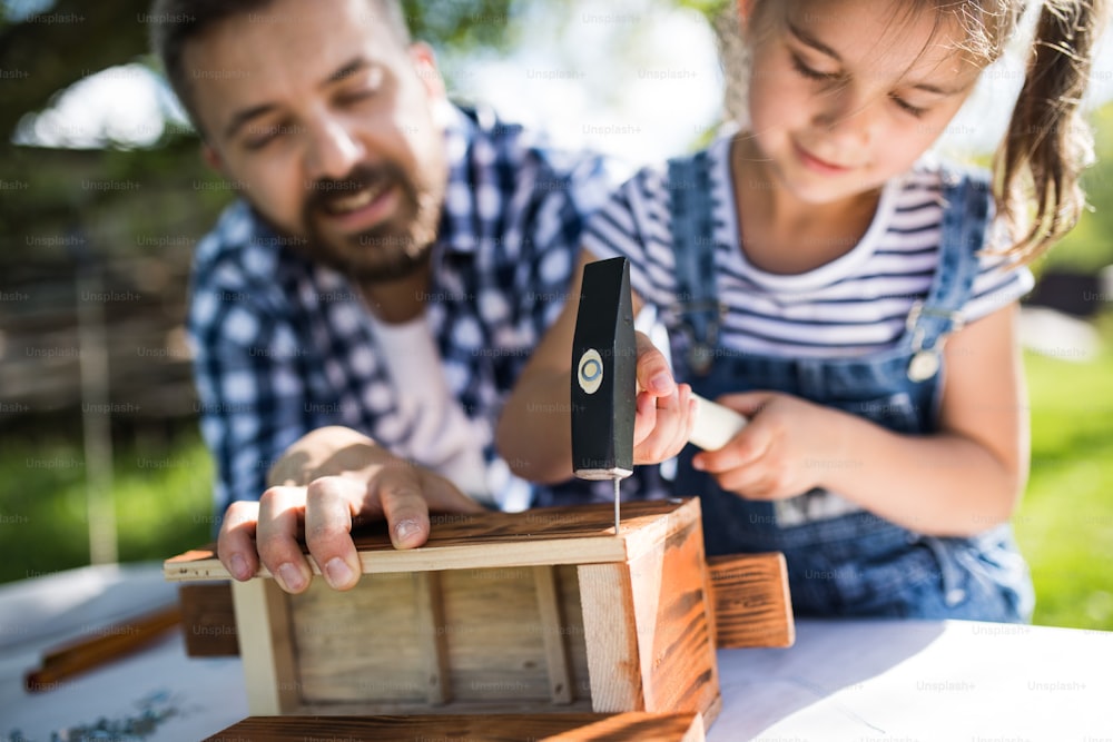 Father with a small daughter outside with hammer making wooden birdhouse or bird feeder.