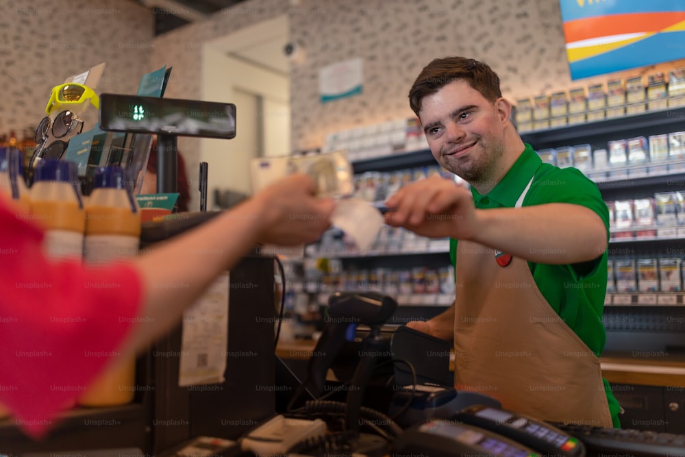 Un joven y alegre empleado con síndrome de Down que recibe el pago en efectivo del cliente en la cafetería de la gasolinera.