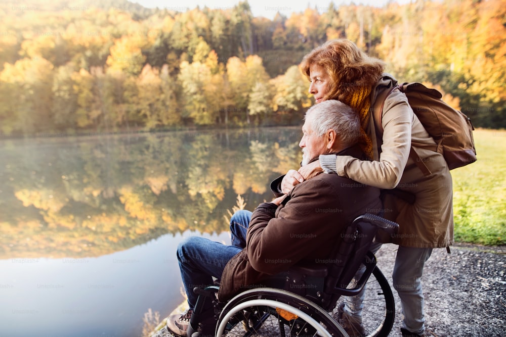 Active senior couple on a walk in a beautiful autumn nature. A woman and man in a wheelchair by the lake in the early morning.