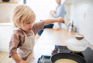 A concentrated small boy helping indoors in kitchen with making pancakes.