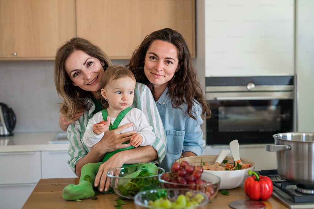 Portrait of mature mother with daughter and granddaughter indoors at home looking at camera when cooking.