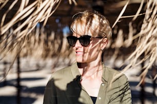 A young woman with sunglasses standing outdoors under straw umbrella on beach.