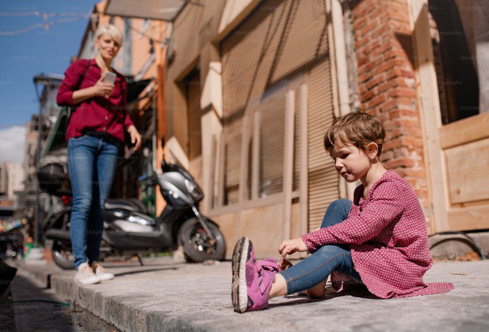 Une petite fille avec une mère assise à l’extérieur sur le trottoir, enlevant ses chaussures.