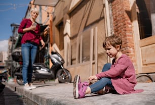 Ein kleines Mädchen mit einer Mutter, die draußen auf dem Bürgersteig sitzt und Schuhe auszieht.