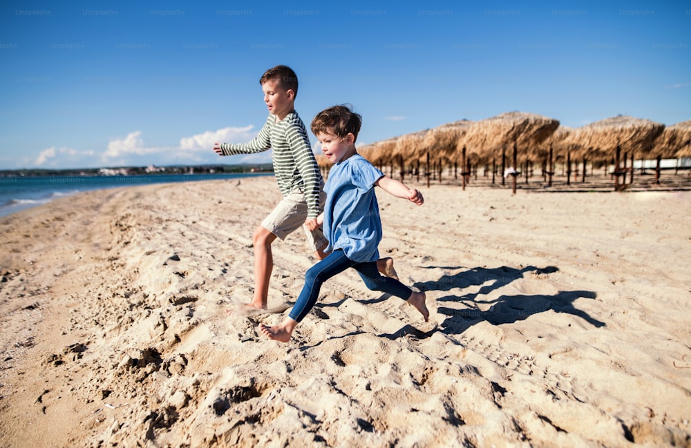 Two small children running outdoors on sand beach, holding hands.