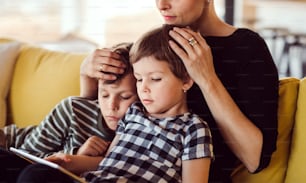 A young unrecognizable woman with two children sitting on sofa indoors at home, using tablet.