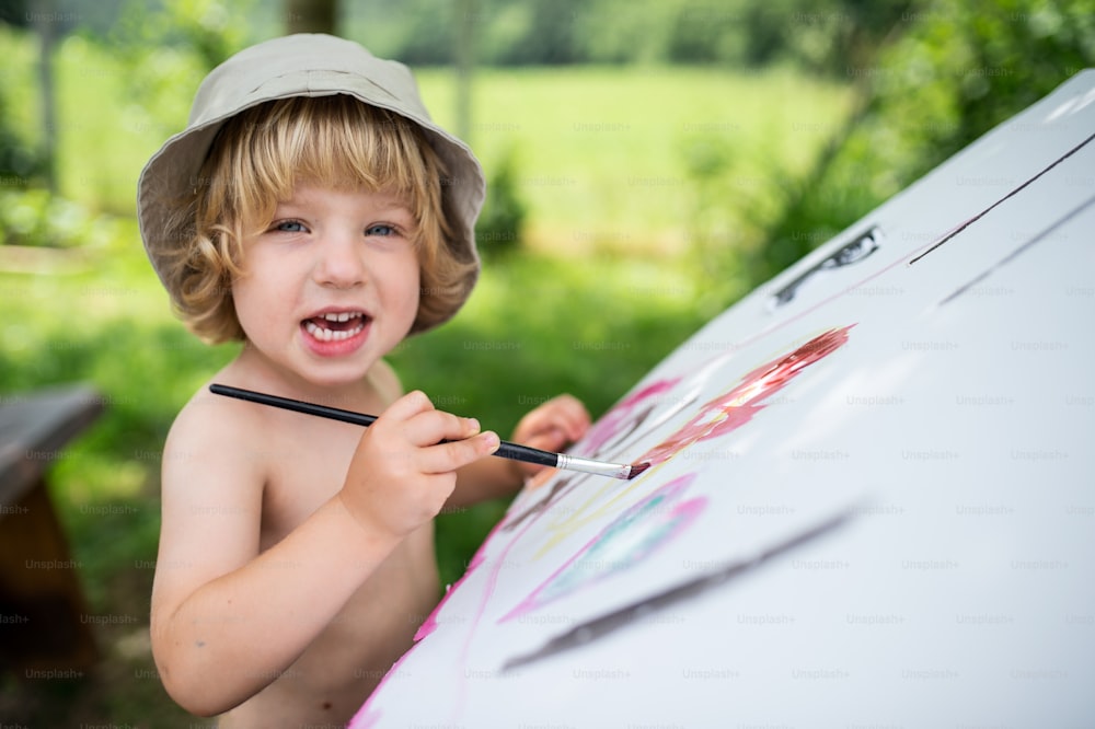 Topless small blond boy with hat painting outdoors in summer, looking at camera.