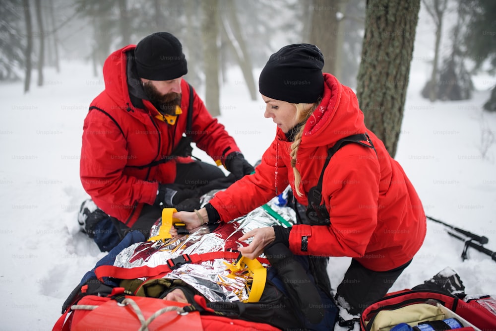 Paramedics from mountain rescue service provide operation outdoors in winter in forest, injured person in stretcher.