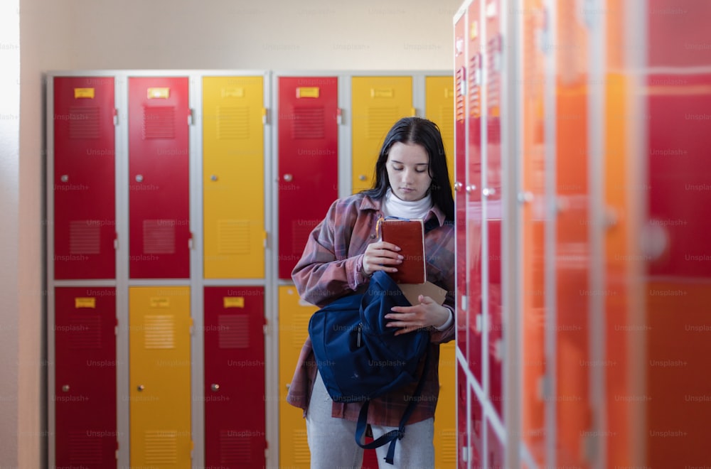 Um estudante adolescente sentado no corredor perto de armários coloridos e embalando livro para mochila no corredor do campus, de volta ao conceito de escola.