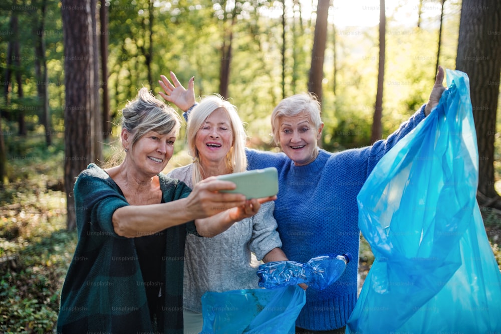 Senior women friends picking up litter outdoors in forest, taking selfie. A plogging concept.