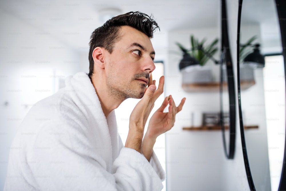 Jeune homme mettant de la crème sur le visage dans la salle de bain le matin, une routine quotidienne.
